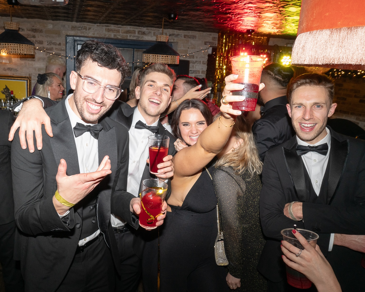 Group of young adults in tuxedos and party dresses holding up vibrant red cocktails and laughing together in a lively bar setting in Bristol.