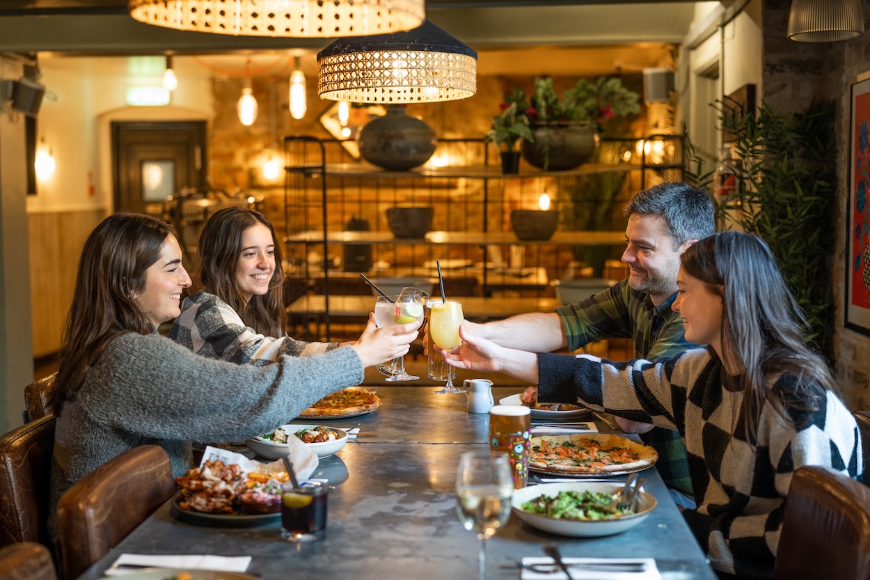 A group of friends toast colourful cocktails over pizza, wings and salad inside the cosy and stylish dining space at Racks—perfect for group catch-ups and private parties in Clifton.